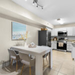 Palm Bay, FL Apartments - Meri Palm Bay - View of Modern Open Kitchen with Stainless Steel Appliances, Granite Counters, White Cabinetry, Cream Tile Floors, and Dining Table with Chairs