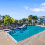 Three-BR Apartments in Palm Bay, FL - Meri Palm Bay - Gated Pool Area with Lounge Chairs, Tables, and Surrounded by Palm Trees